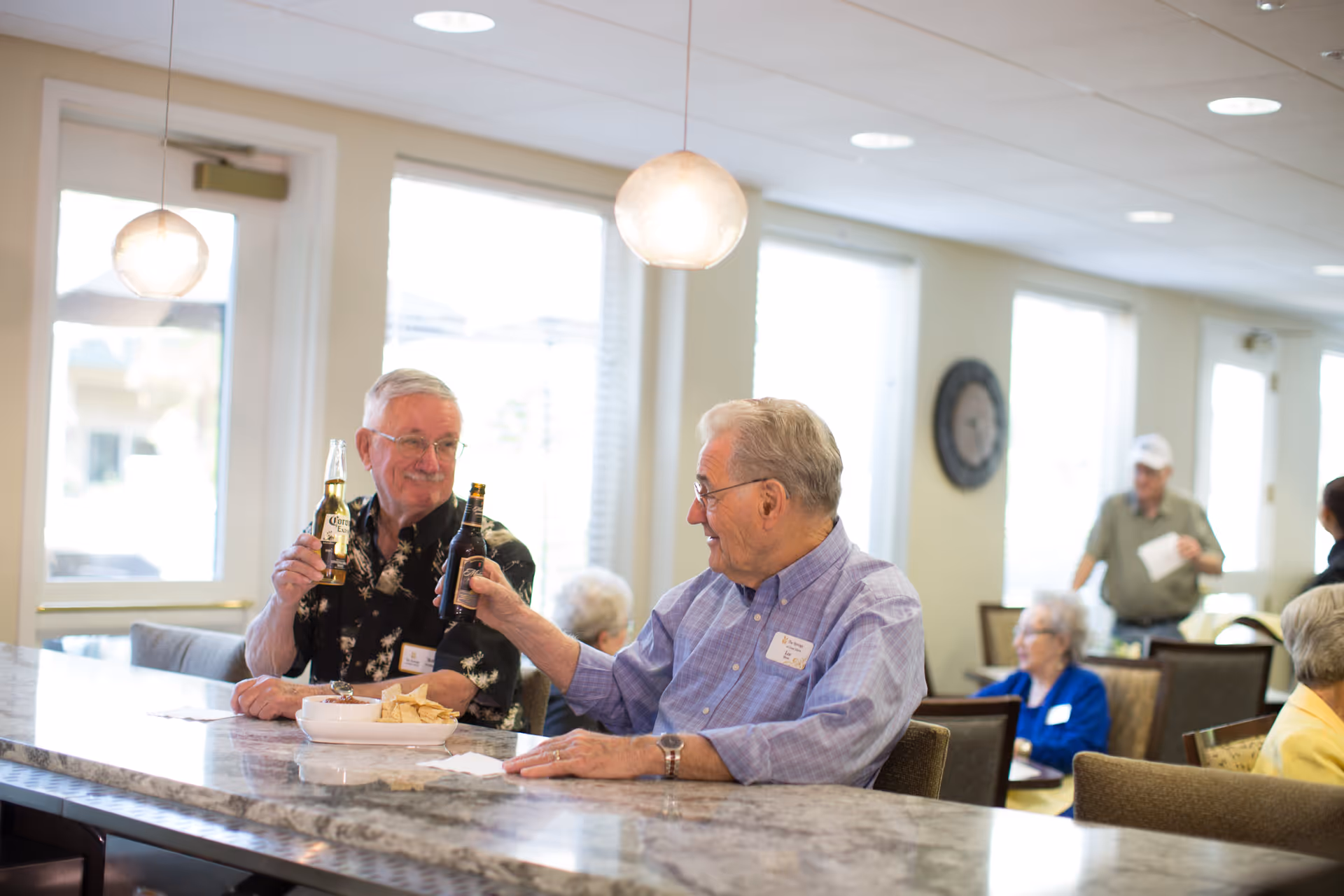 Two elderly men sitting at a marble countertop in a well-lit room, clinking their beer bottles together in a toast. There are snacks on the counter and other elderly people sitting and standing in the background.