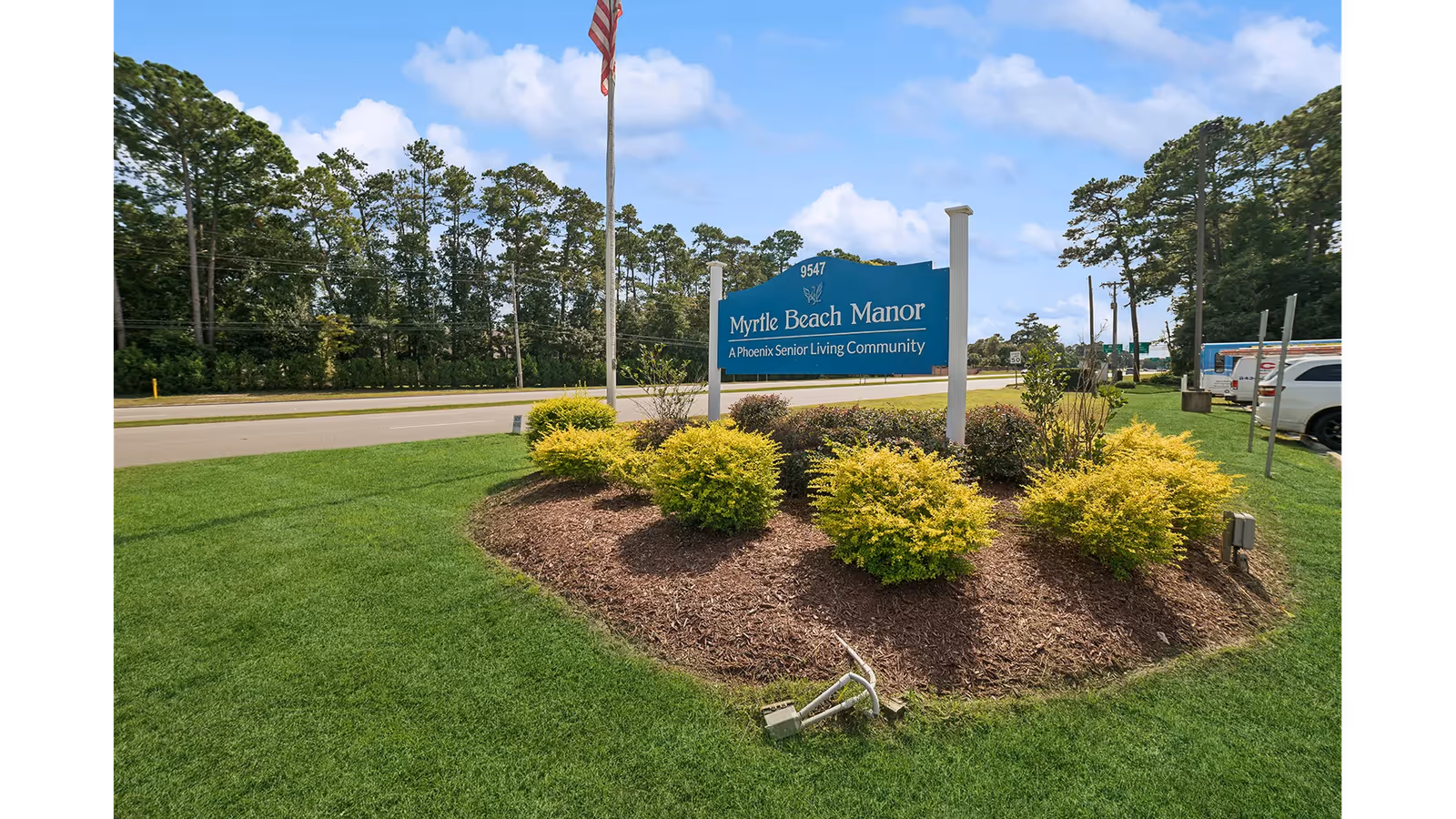 Entrance sign for Myrtle Beach Manor surrounded by landscaped shrubs and an American flag near a roadside.