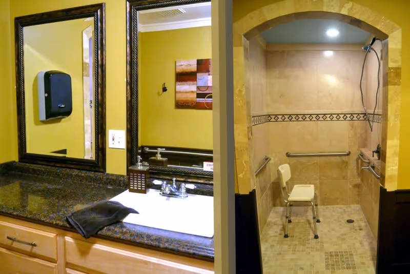 A bathroom area featuring a granite countertop with a sink, a dark towel, and a soap dispenser. Above the sink is a large framed mirror reflecting part of the room. To the right, there is a walk-in shower with beige tiles, a built-in bench, grab bars, and a handheld showerhead, all under a recessed light.