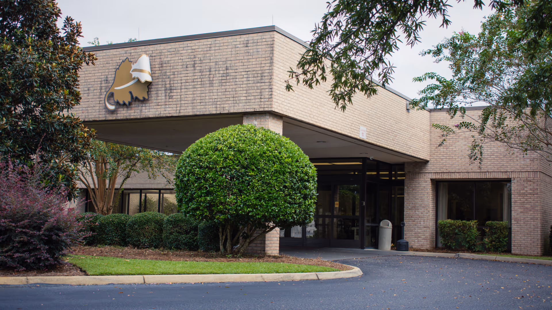 Exterior view of a single-story brick building with a covered entrance, surrounded by neatly trimmed bushes and trees under a cloudy sky.