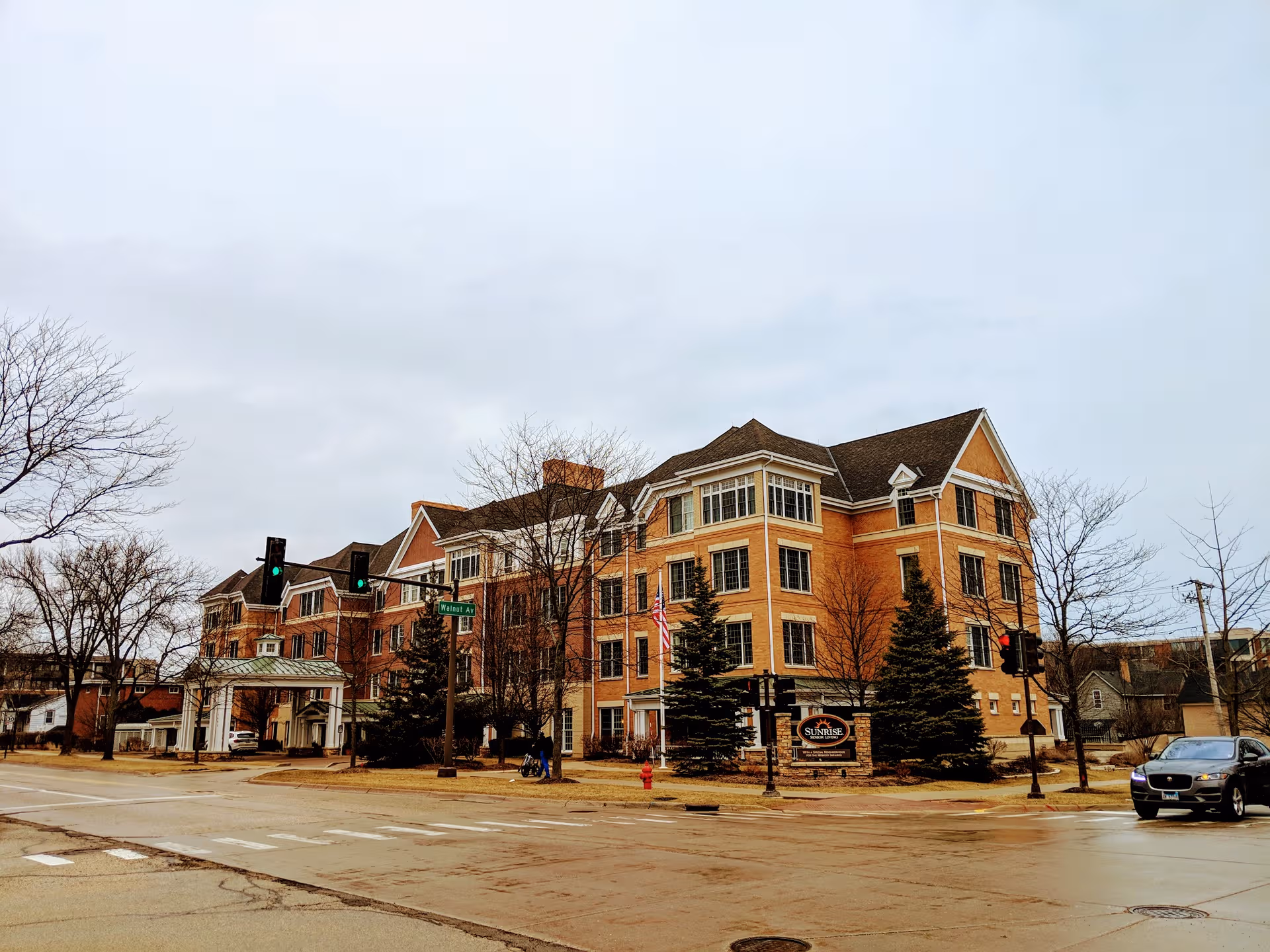 Brick senior living building on a street corner with trees and a car