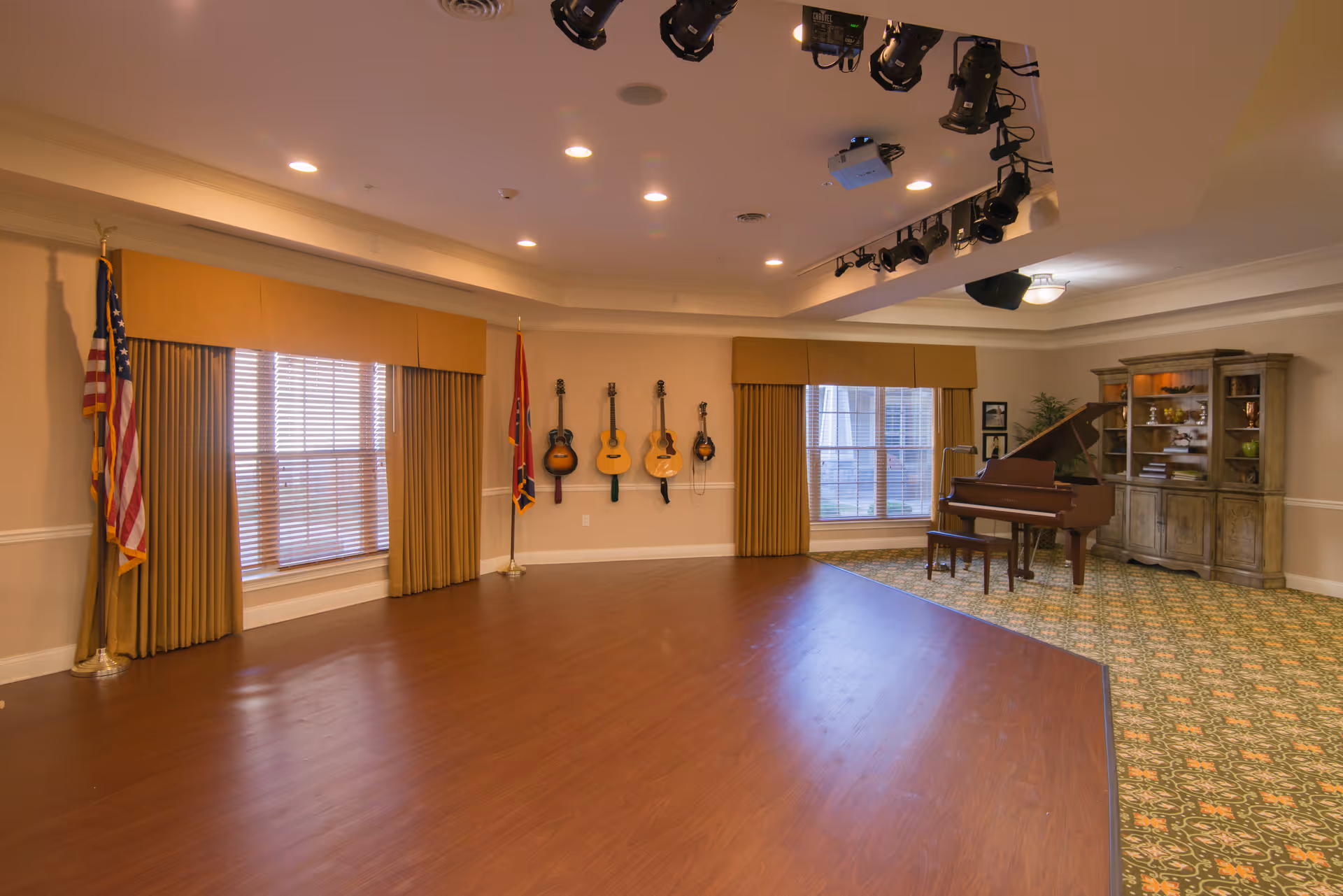 Large community activity room with a small stage, grand piano, wall-mounted guitars, flags, and a display cabinet.