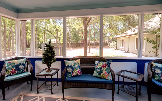 A sunroom seating area features wicker chairs, colorful cushions, and large windows overlooking trees.