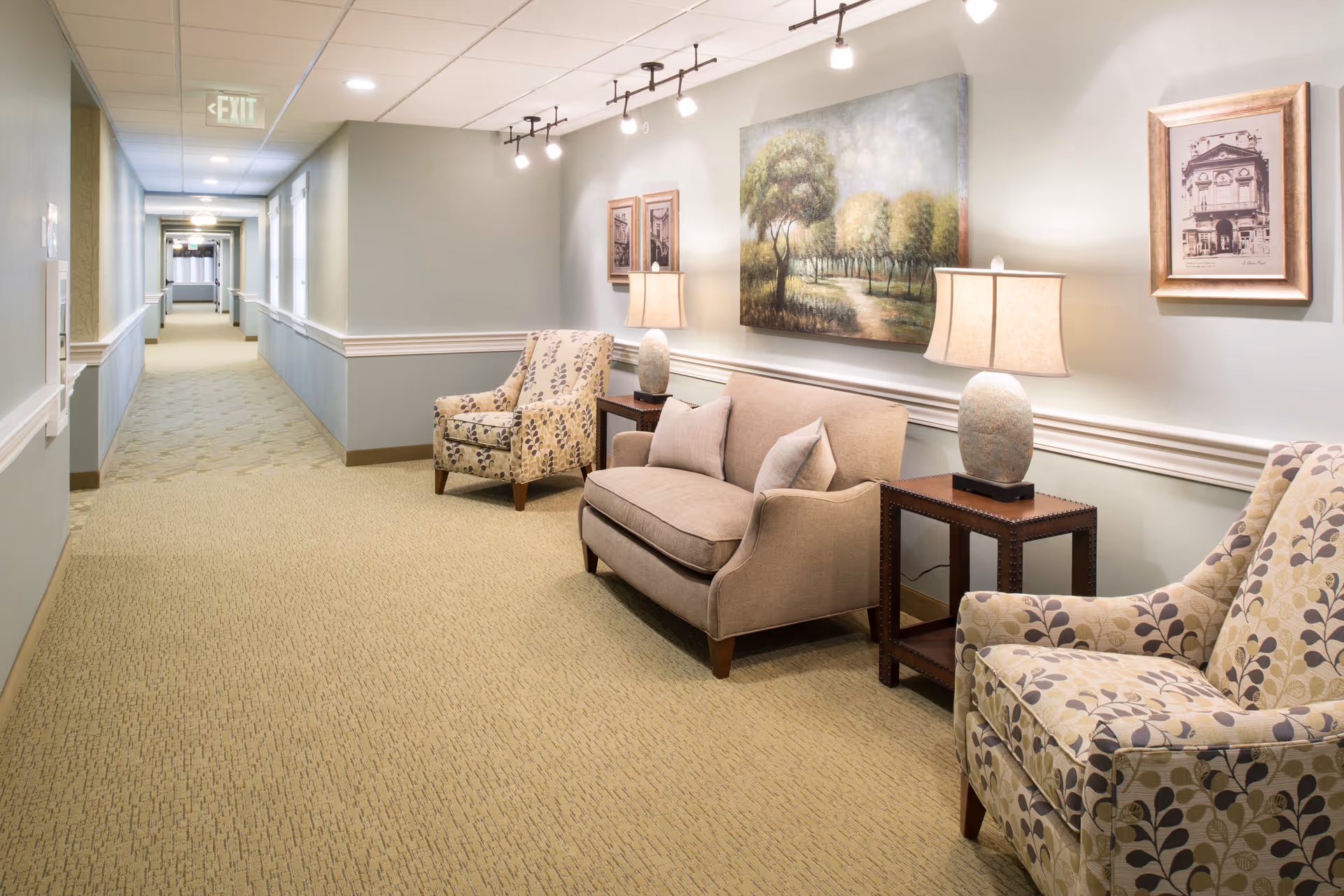 Well-lit hallway seating area with upholstered chairs, a loveseat, side tables, lamps and wall art.
