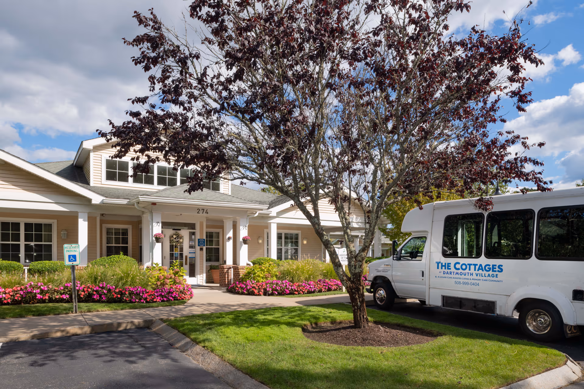 Front entrance of a cottage-style senior living building with landscaped flower beds and a shuttle van parked outside.