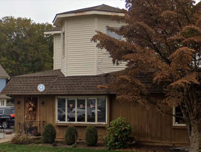 Front view of a brown single-story building with a white two-story addition and trees.