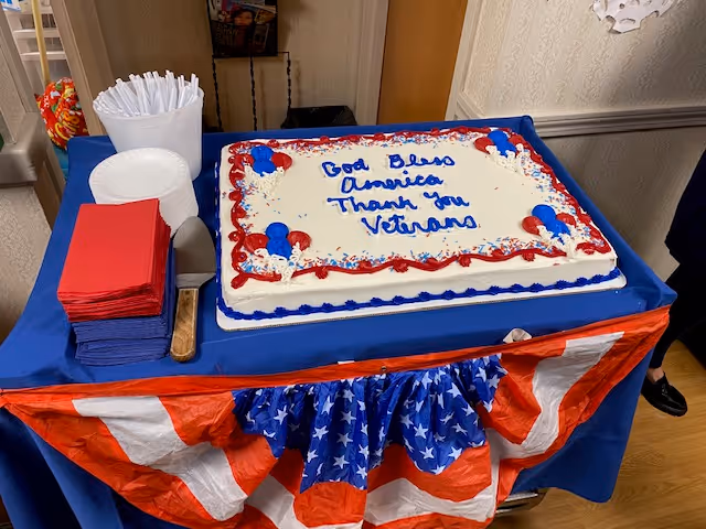 A rectangular cake decorated with red, white, and blue icing on a table covered with a blue cloth and patriotic red, white, and blue bunting. The cake has the message 'God Bless America Thank You Veterans' written in blue icing. Next to the cake are stacks of red and blue napkins, white disposable plates, and a container holding white plastic forks.