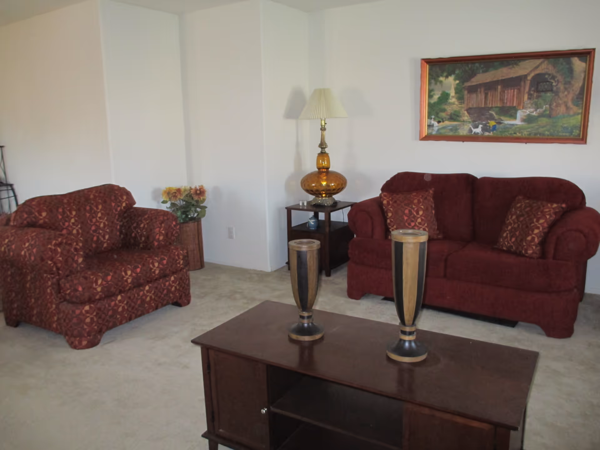 A living room with a red patterned armchair and a matching red loveseat with two patterned cushions. There is a wooden coffee table with two decorative vases on it in the foreground. A small side table with a large amber glass lamp and a framed painting of a covered bridge and nature scene hangs on the white wall behind the loveseat. A basket with artificial sunflowers is placed in the corner.