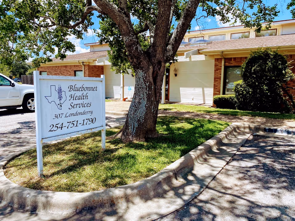 Exterior of Bluebonnet Health Services Hospice building with a large sign and tree.