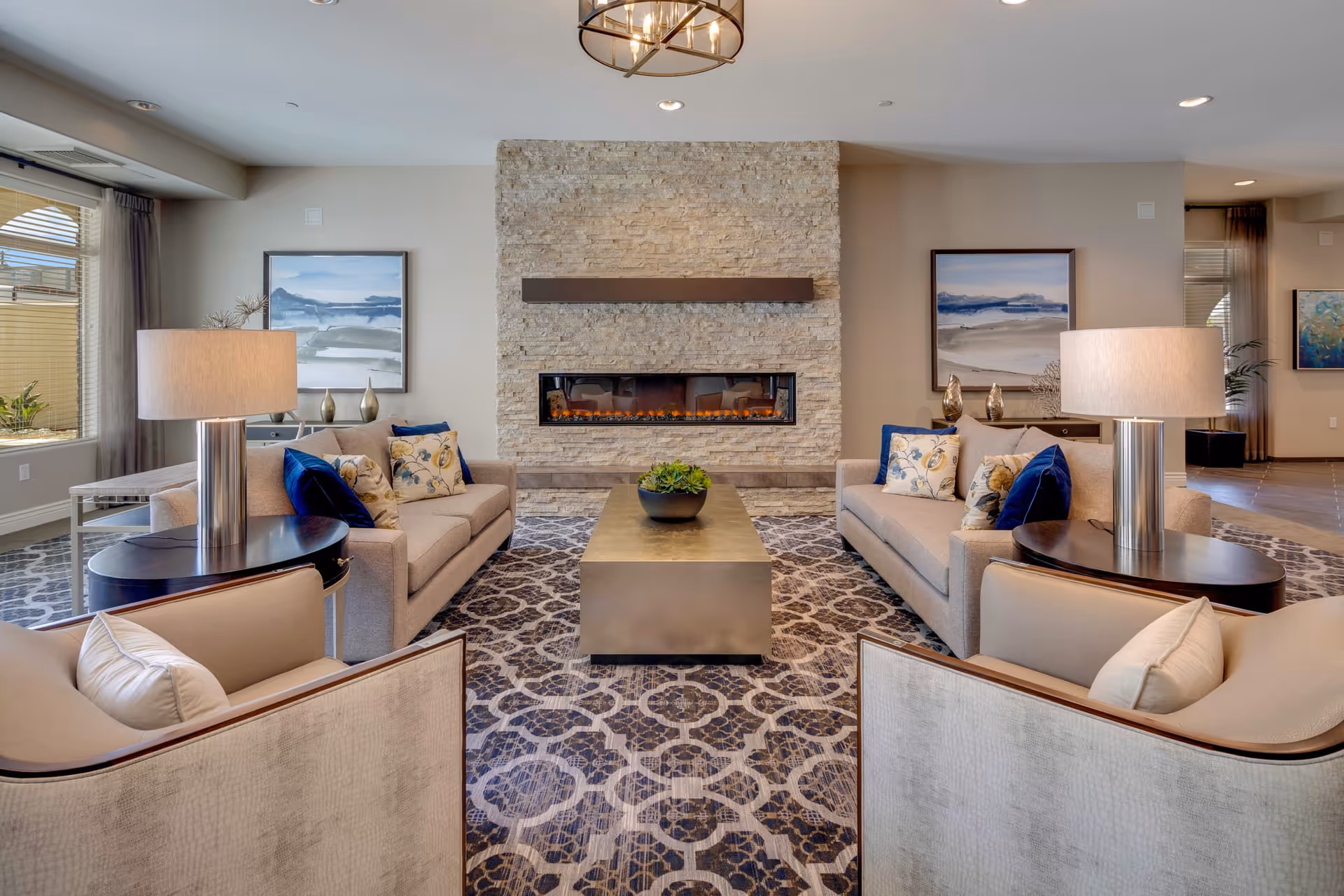 A cozy living room area featuring two beige sofas and two armchairs arranged around a rectangular coffee table. The room has a patterned carpet, two table lamps on side tables, and a modern stone fireplace centered on the far wall. Two framed landscape paintings hang on the walls beside the fireplace, and large windows with curtains allow natural light to fill the space.