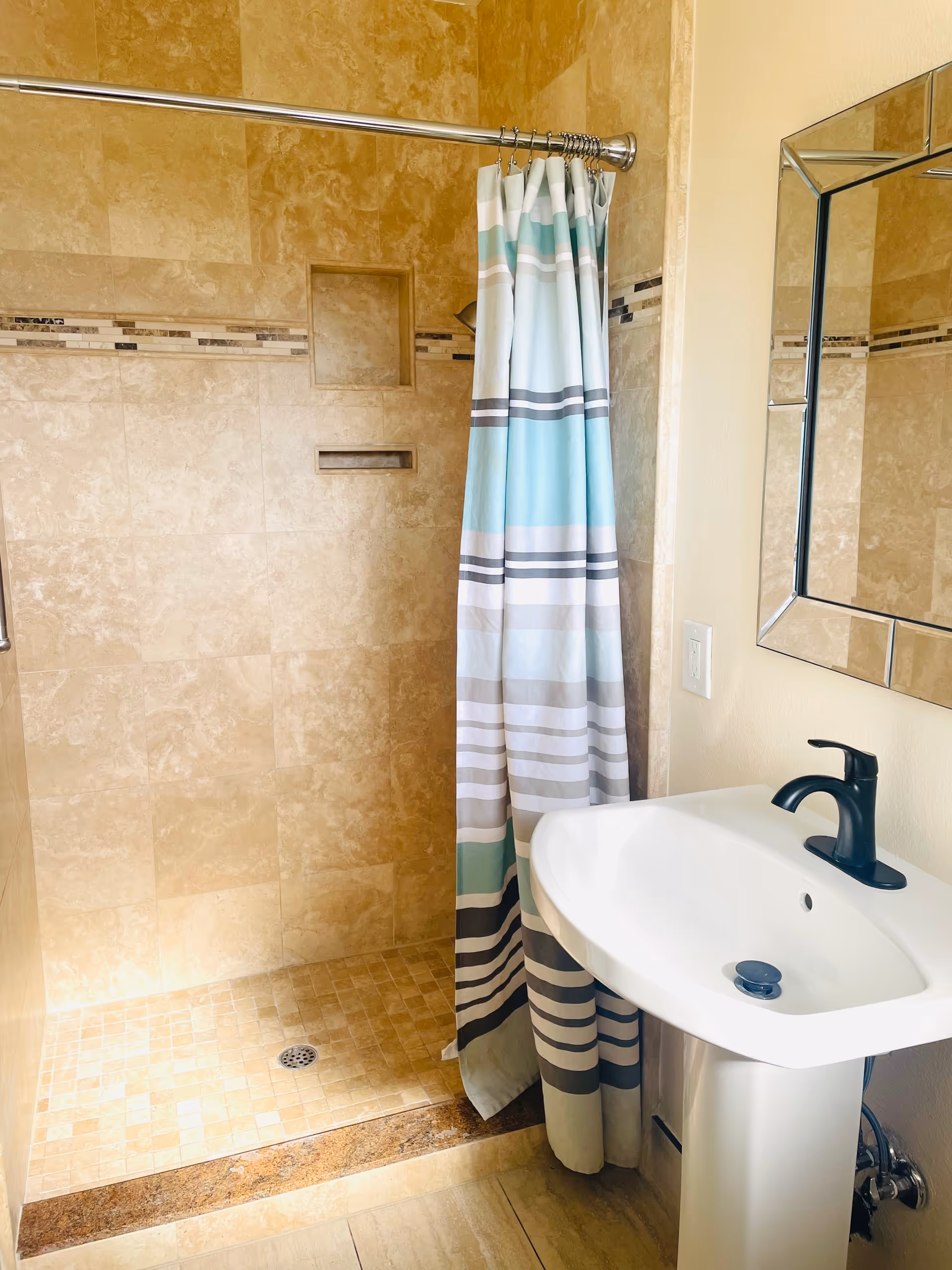 A bathroom with beige tiled walls and floor featuring a walk-in shower with a striped shower curtain in shades of blue, gray, and white. There is a white pedestal sink with a black faucet and a rectangular mirror with a decorative frame mounted on the wall above the sink.