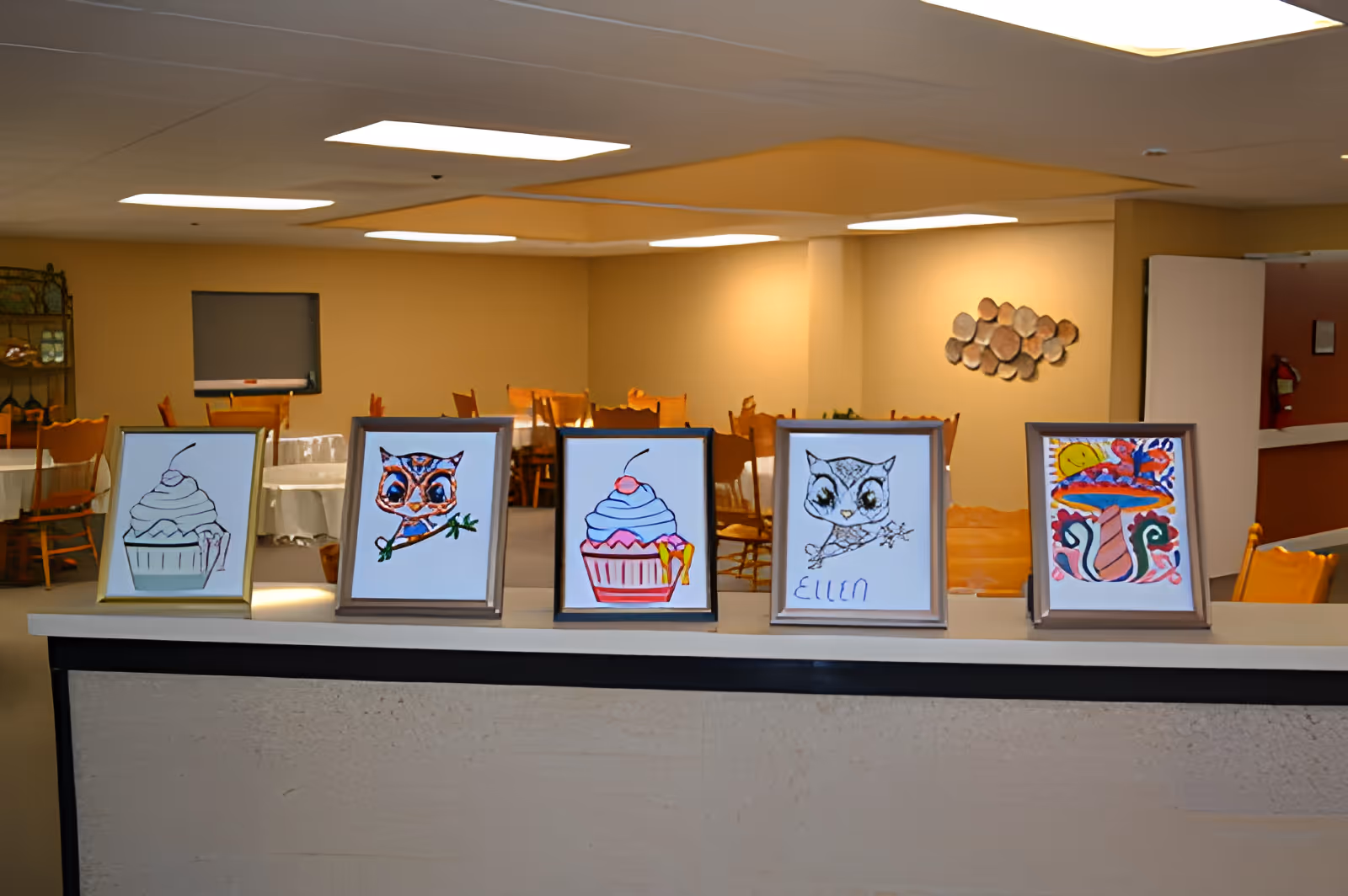 Five framed colorful drawings displayed on a counter with a dining area and tables and chairs in the background.
