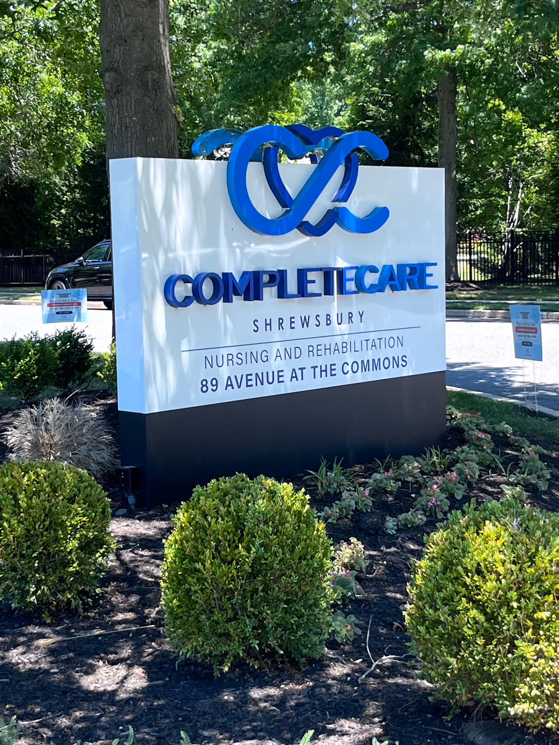 A white and blue Complete Care Shrewsbury sign outdoors among landscaped bushes.