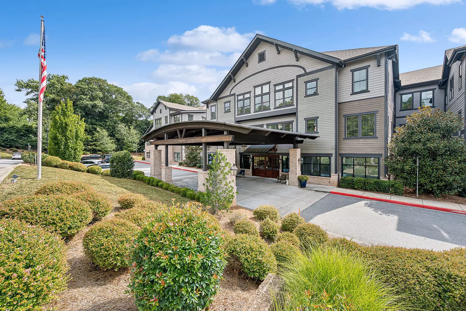 Front entrance of a multi-story senior living facility with a covered porte-cochere, landscaped shrubs, and an American flag.
