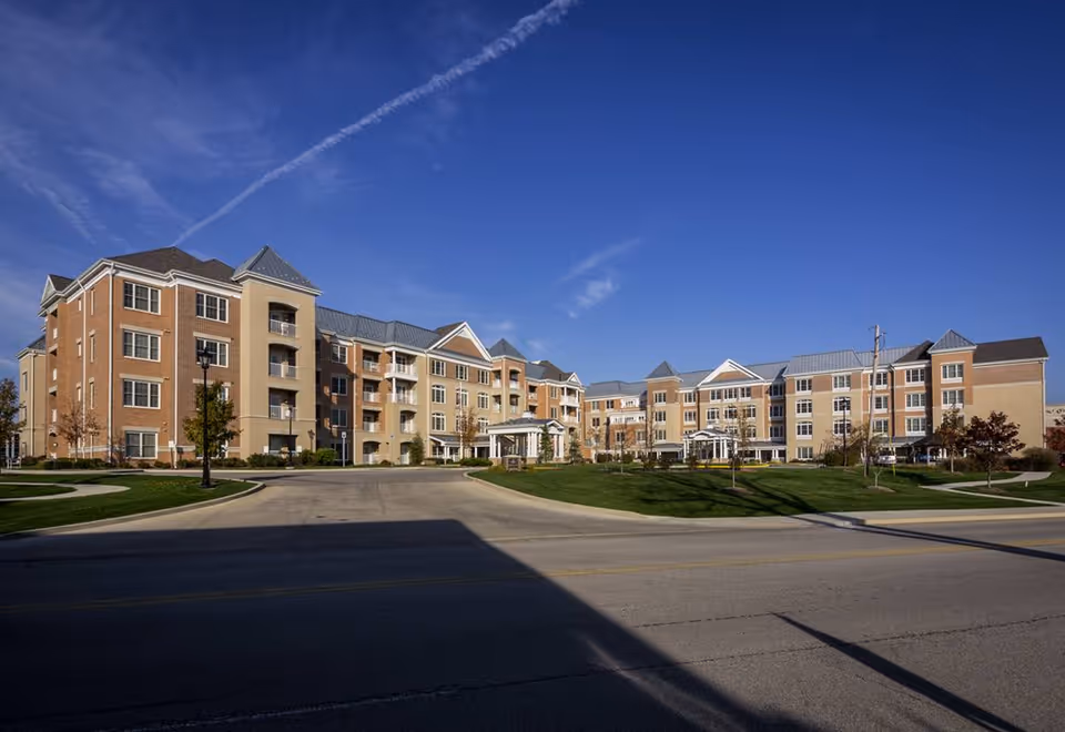 Large four-story senior living building with brick and beige facade, front driveway and landscaped lawn under a clear blue sky.