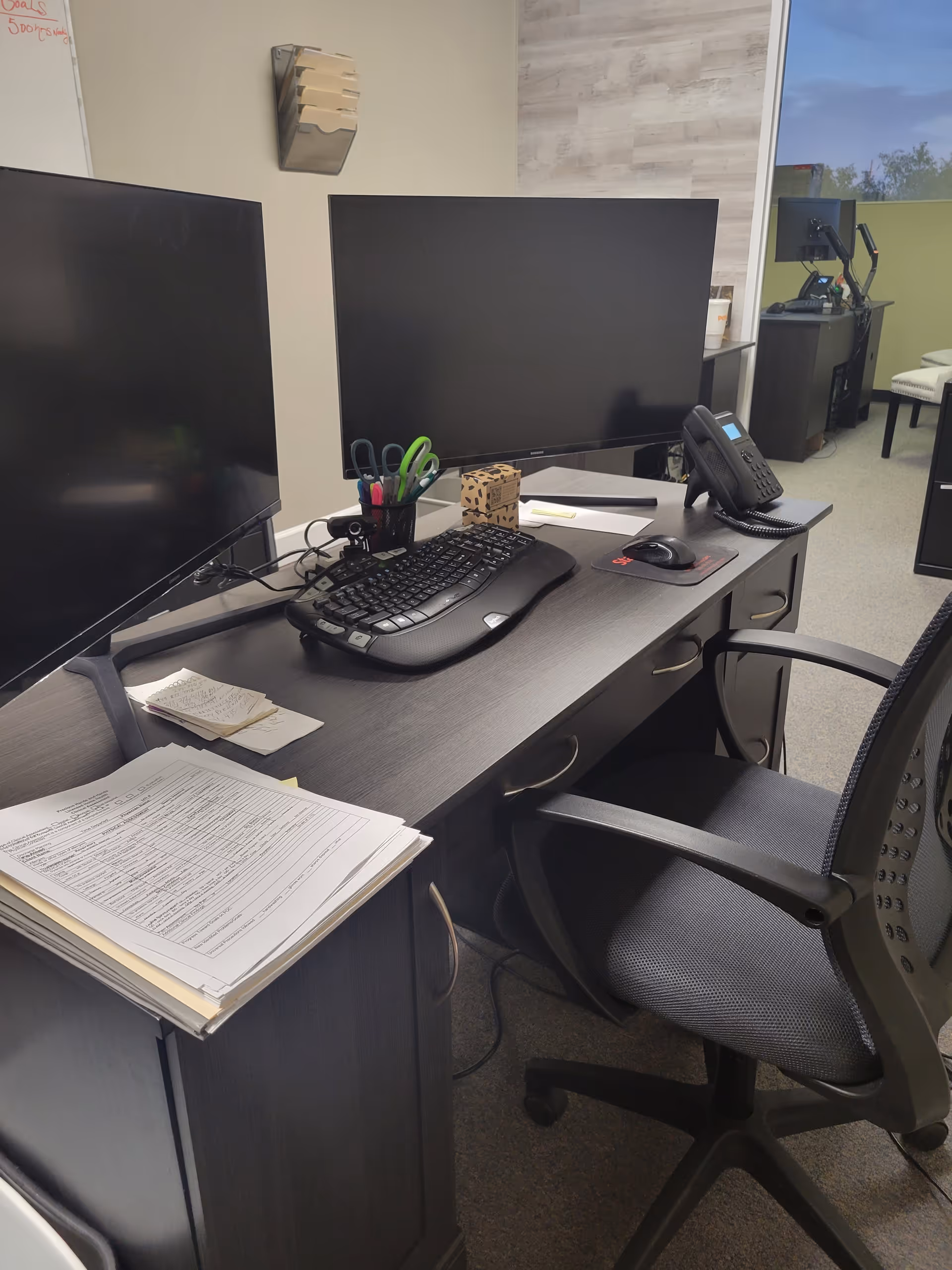 Office desk with dual monitors, keyboard, mouse, phone, and scattered paperwork.