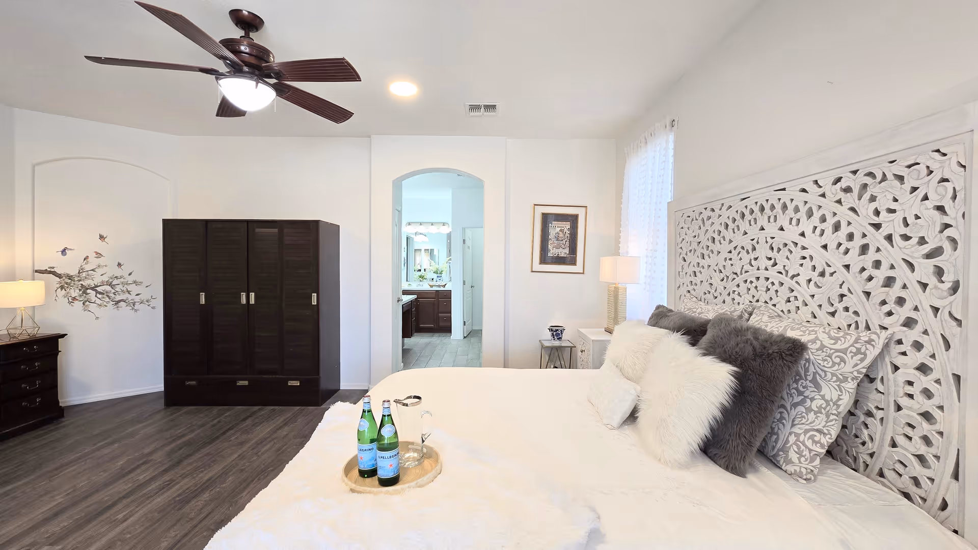 Spacious bedroom features ornate carved headboard, dark wardrobe, ceiling fan, and tray of beverages on bed.