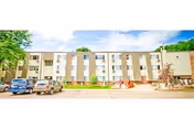 Three-story beige senior living building with cars parked in front and a landscaped entrance.