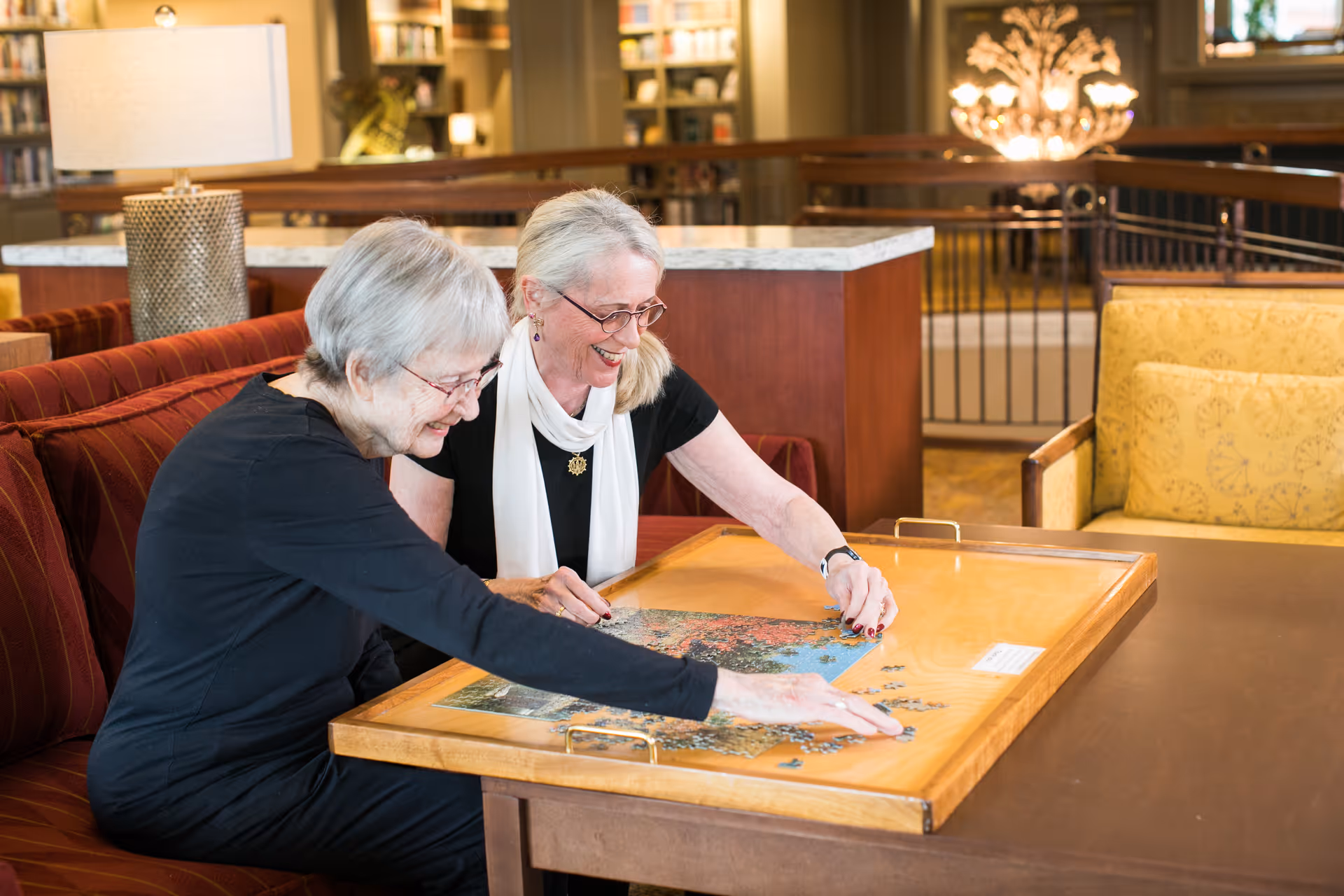 Two elderly women sitting on a red couch in a well-lit room, smiling and working together on assembling a jigsaw puzzle on a wooden tray table. The background shows bookshelves and a lamp, suggesting a cozy common area.