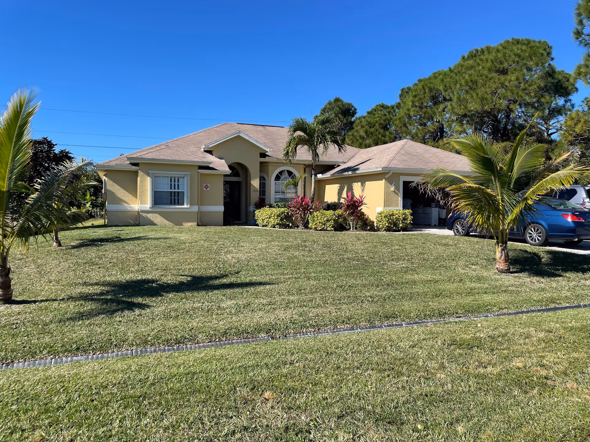 Single-story yellow house with a manicured front lawn, palm trees, and an attached garage with cars in the driveway.
