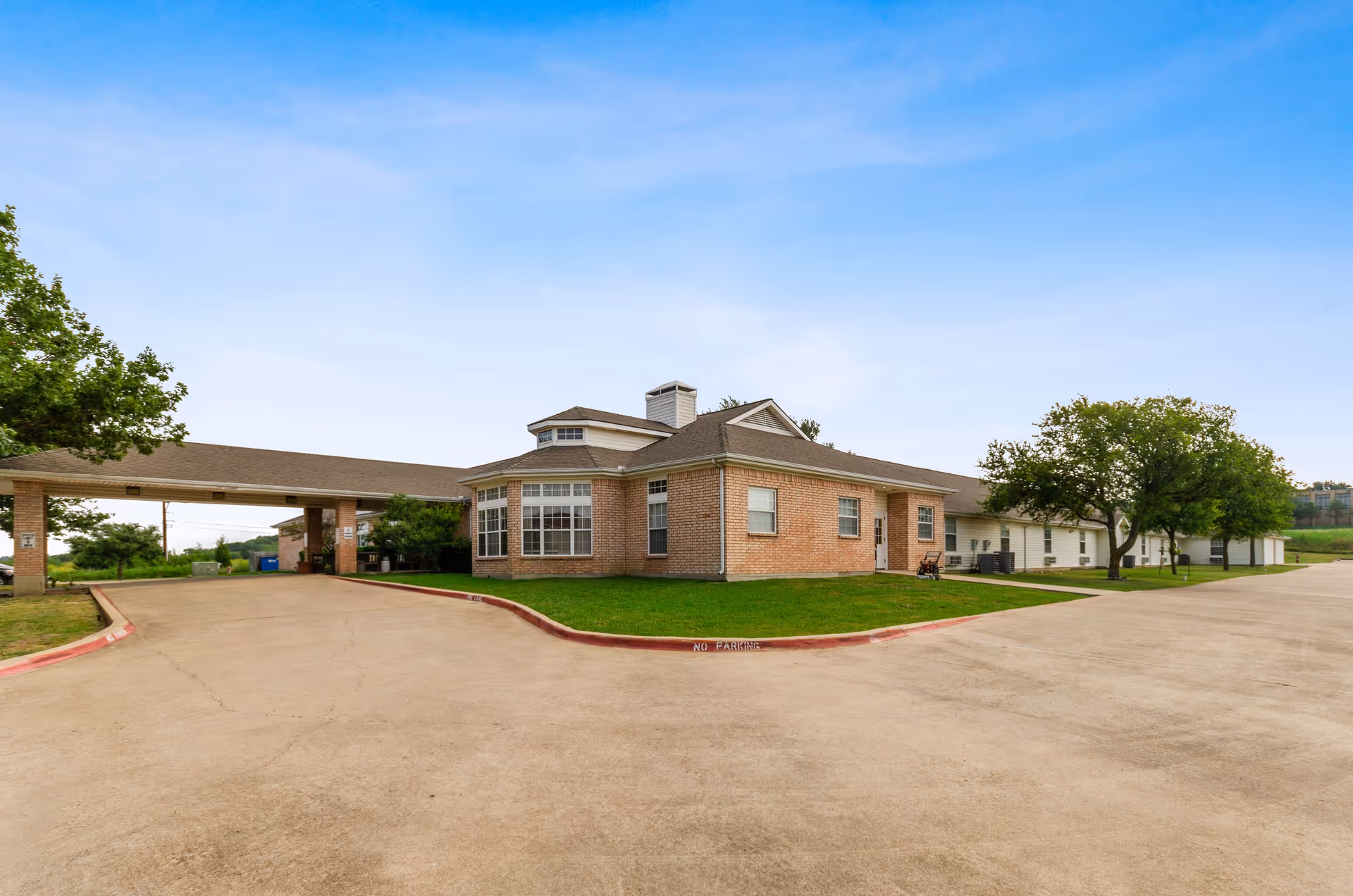 Low-rise brick assisted living building with a covered drive-through entrance, lawn, trees, and a wide paved parking area under a blue sky.