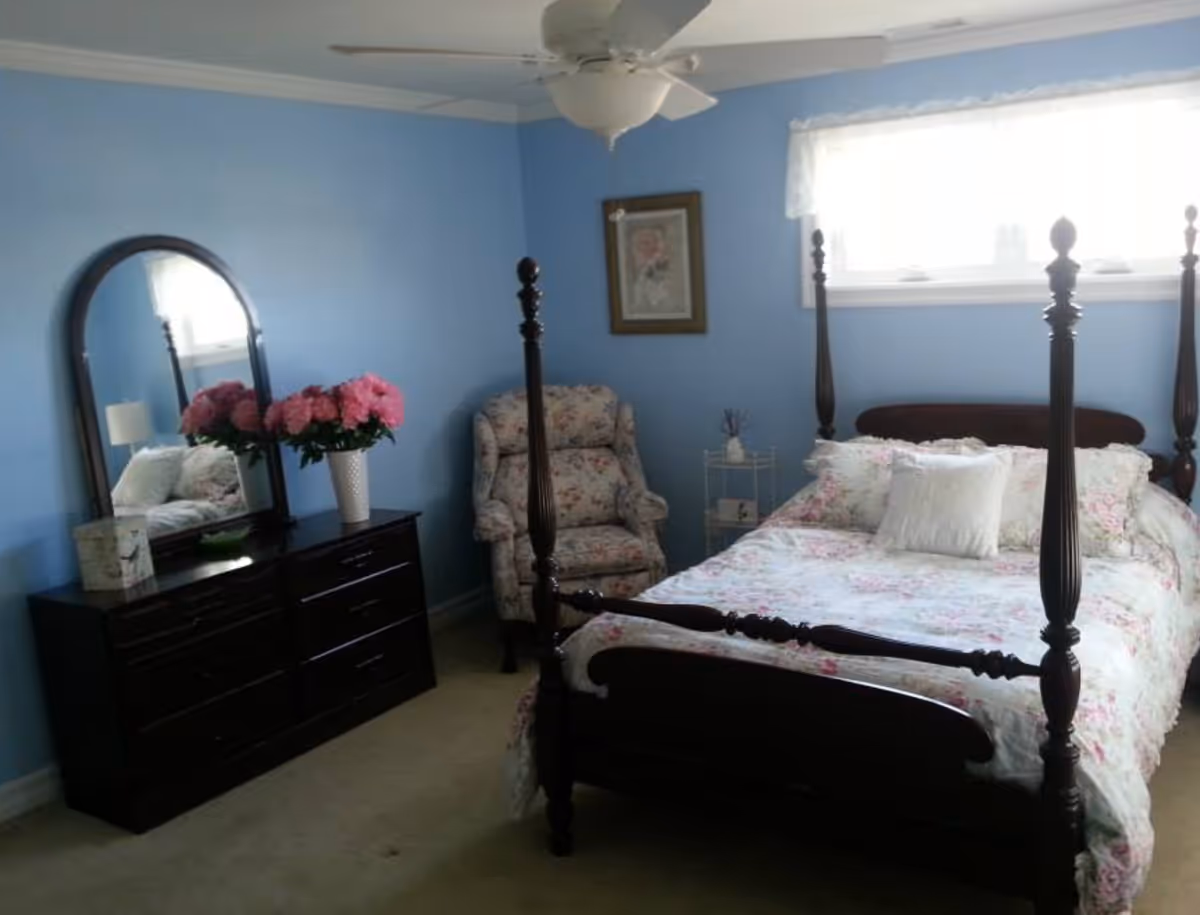 Sunlit bedroom with a dark wood four-poster bed, floral bedding, dresser with mirror and vase of pink flowers against light blue walls.