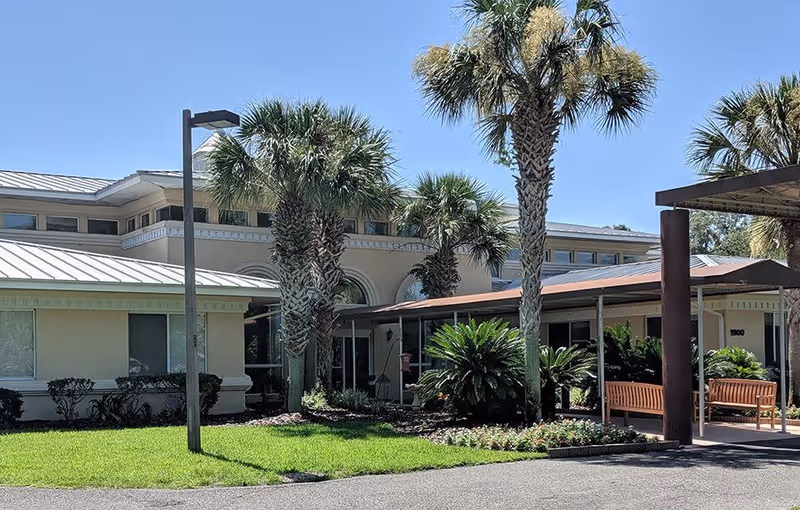 Front entrance of a low-rise beige building with palm trees, a covered walkway and benches.