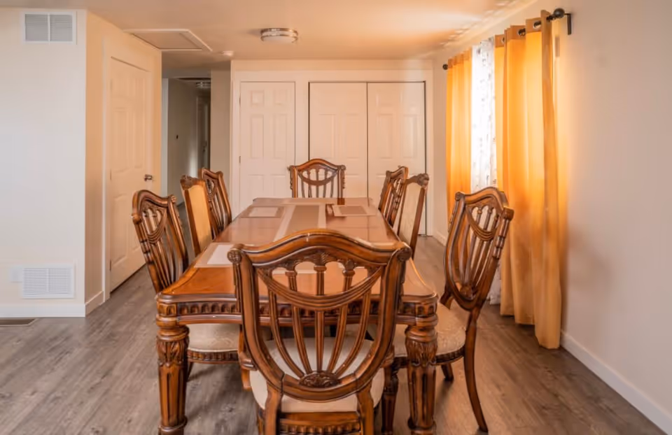 A dining room with a wooden dining table and six matching chairs. The room has light-colored walls, wood flooring, and a window with yellow curtains on the right side. There are two closed white doors at the back of the room.