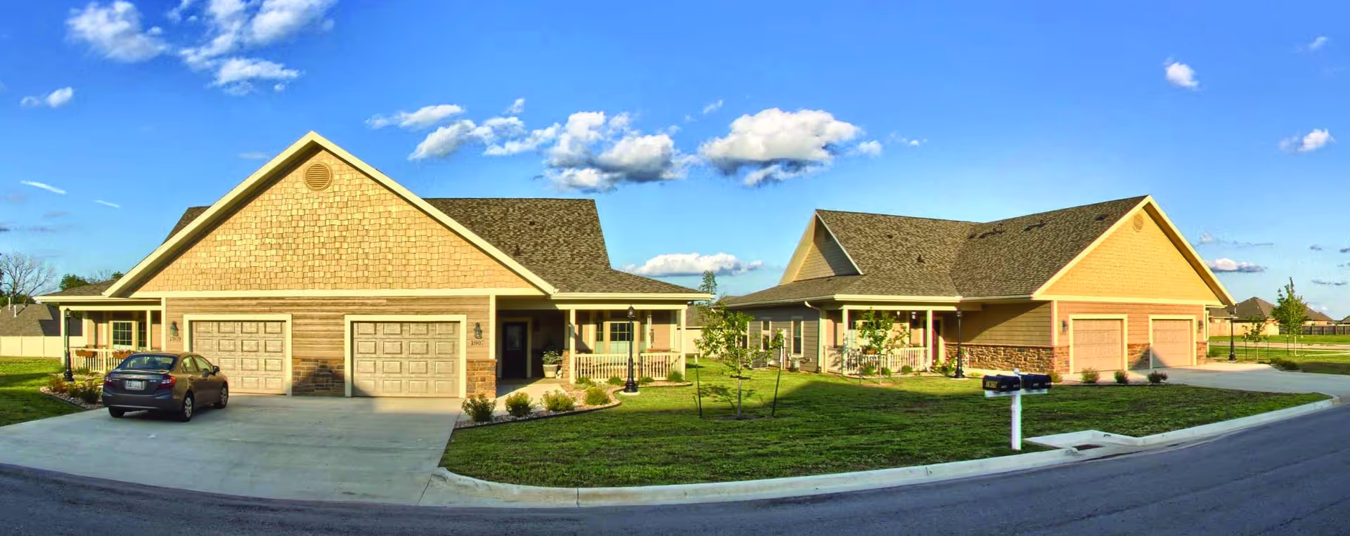 Exterior view of two single-story residential buildings with garages, driveways, and well-maintained lawns under a blue sky with scattered clouds.