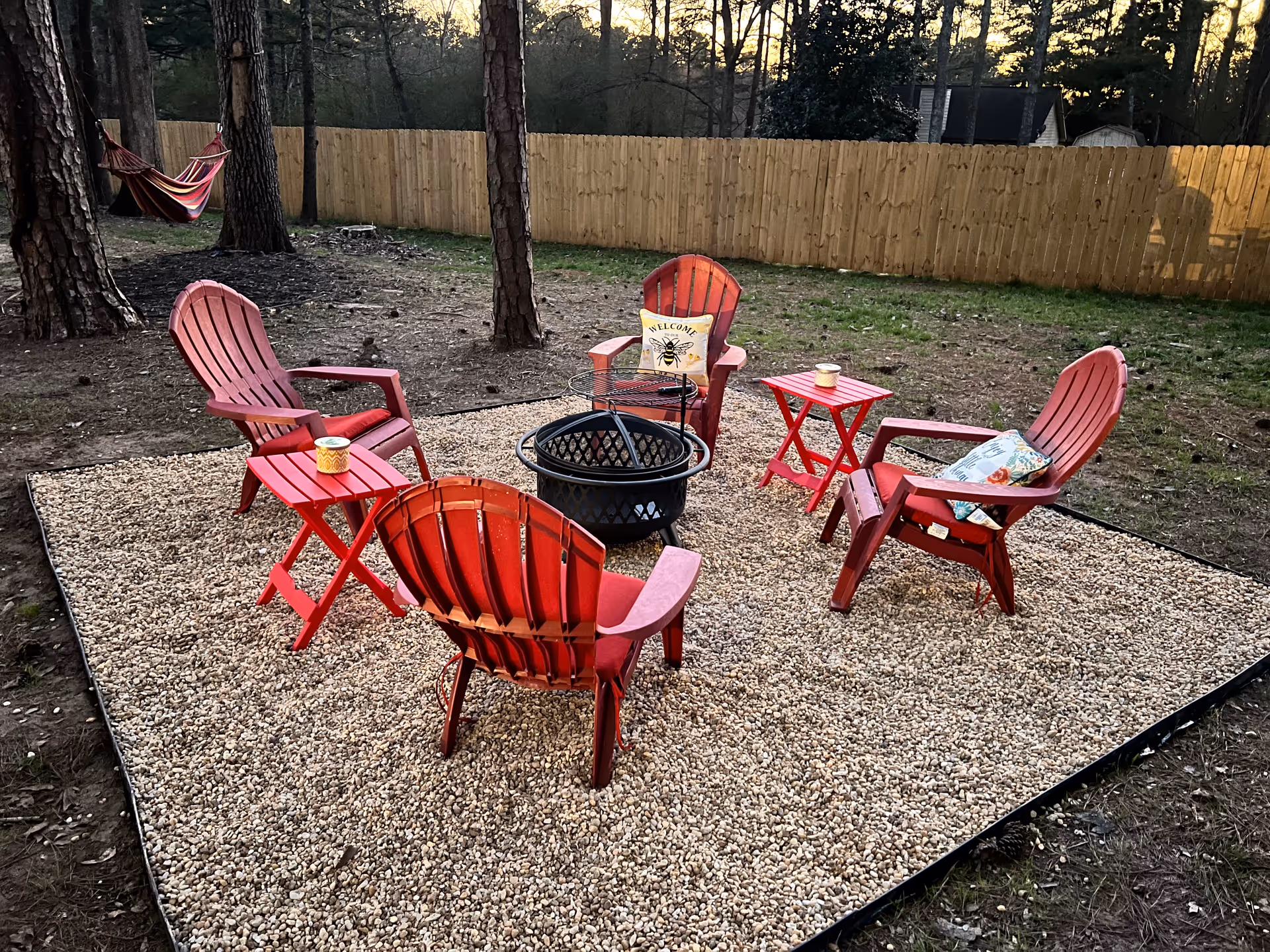 A circular arrangement of red Adirondack chairs around a fire pit on a gravel patio outdoors.