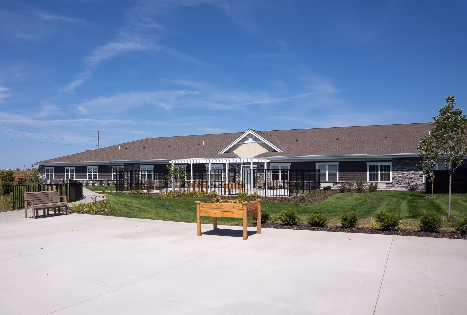 Single-story senior living facility building with a brown roof, stone and dark siding exterior, surrounded by a well-maintained lawn, small shrubs, a wooden bench, and a raised garden bed under a clear blue sky.