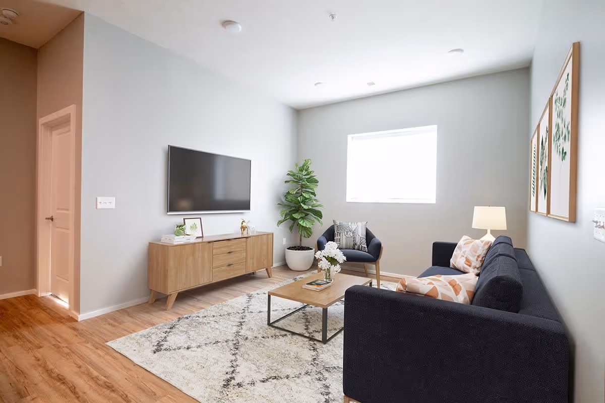 A modern living room with a dark blue sofa and armchair, a wooden coffee table with a vase of flowers, a patterned rug, a wooden TV stand with decorative items, a flat-screen TV mounted on the wall, a large potted plant, and three framed botanical prints on the wall.