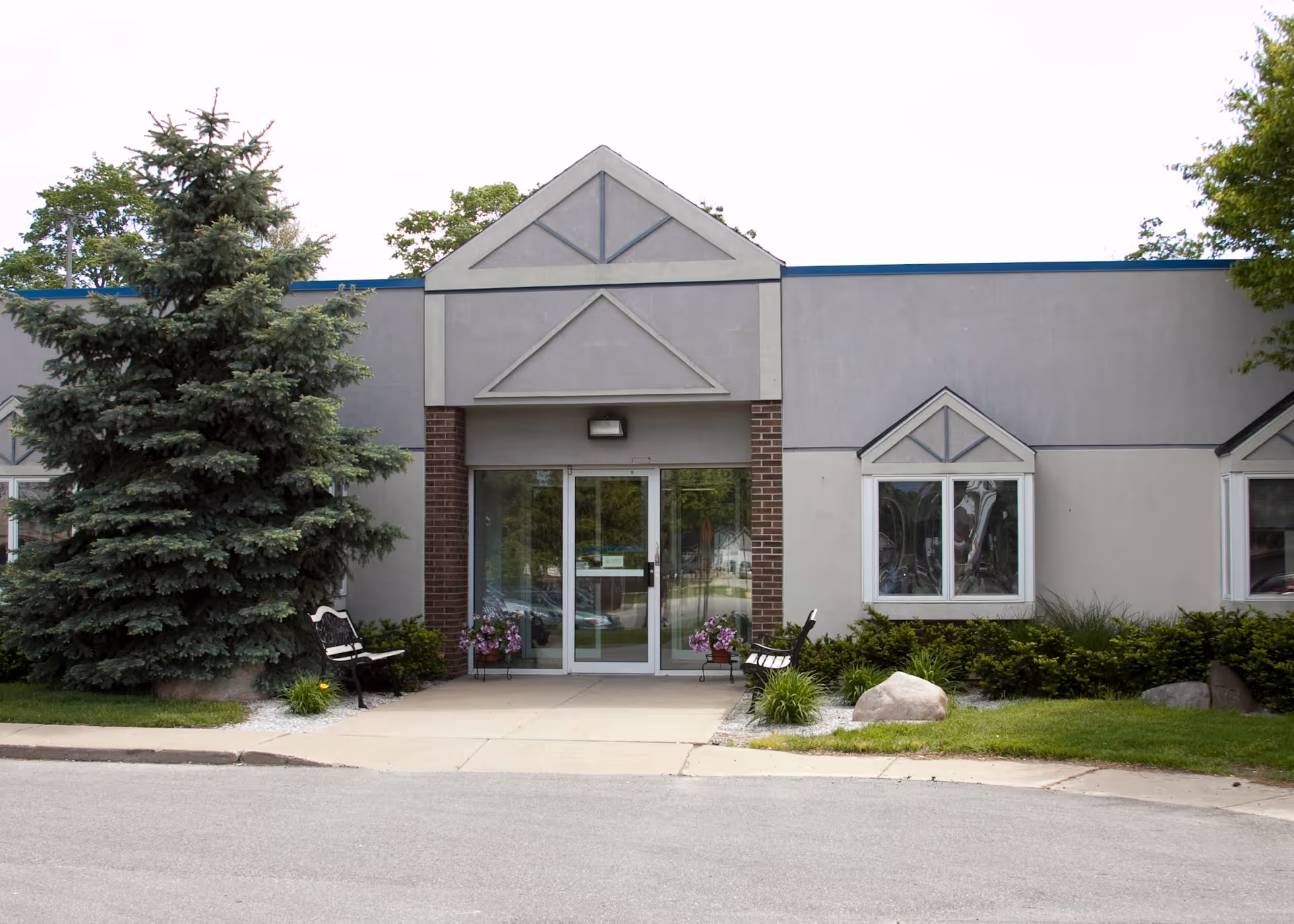 Front exterior view of Fountainview Retirement Village of Grant building with a glass entrance door, two benches on either side, flower pots, and landscaping including a large evergreen tree and bushes.