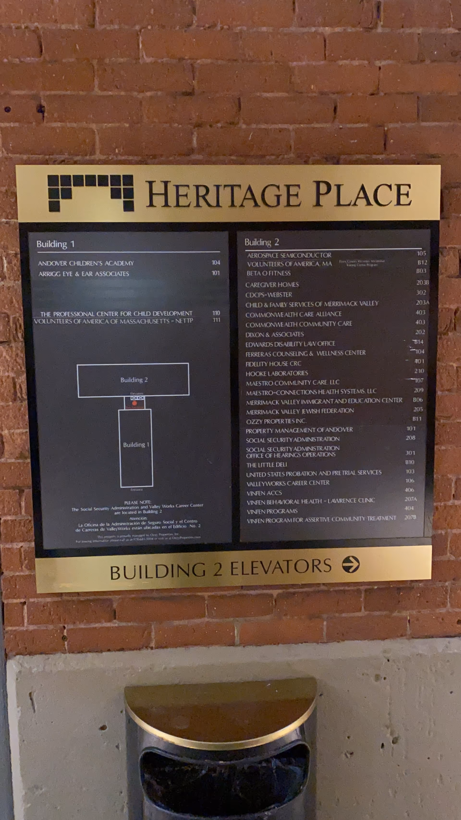 A brass Heritage Place directory sign on a brick wall with building map and trash bin.