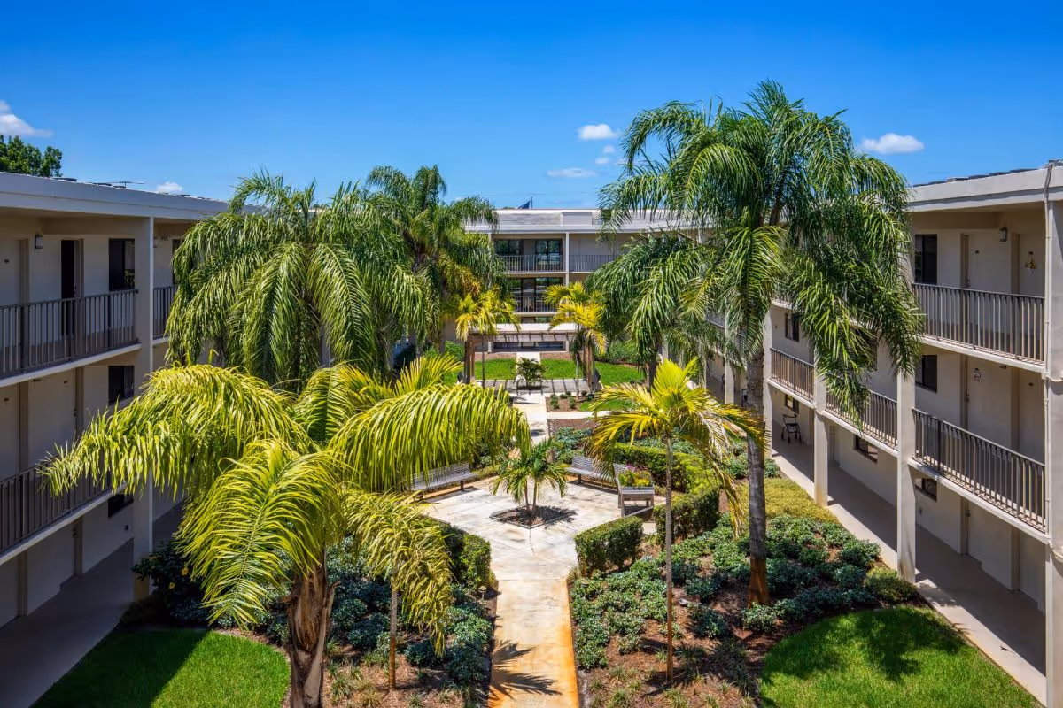 View of a courtyard garden area with palm trees and landscaped greenery, surrounded by a two-story building with balconies and walkways under a clear blue sky.