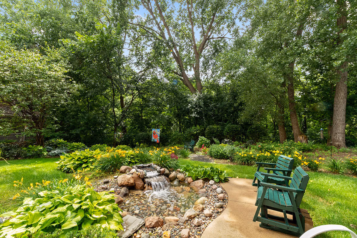 A peaceful garden area with lush green trees and plants surrounding a small rock-lined pond with a waterfall. There are two green wooden benches on a curved concrete path beside the pond, and colorful flowers are blooming throughout the garden.