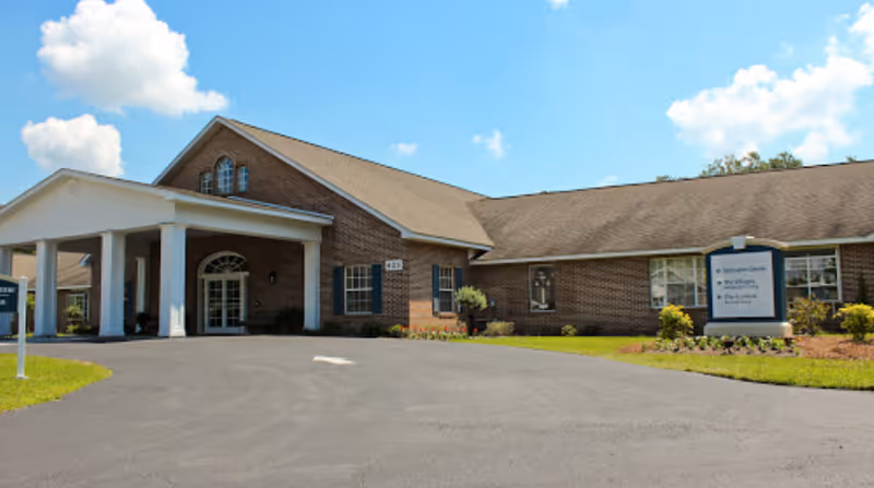 Exterior view of a single-story brick building with a covered entrance supported by white columns, surrounded by a paved driveway and landscaped greenery under a blue sky with some clouds.