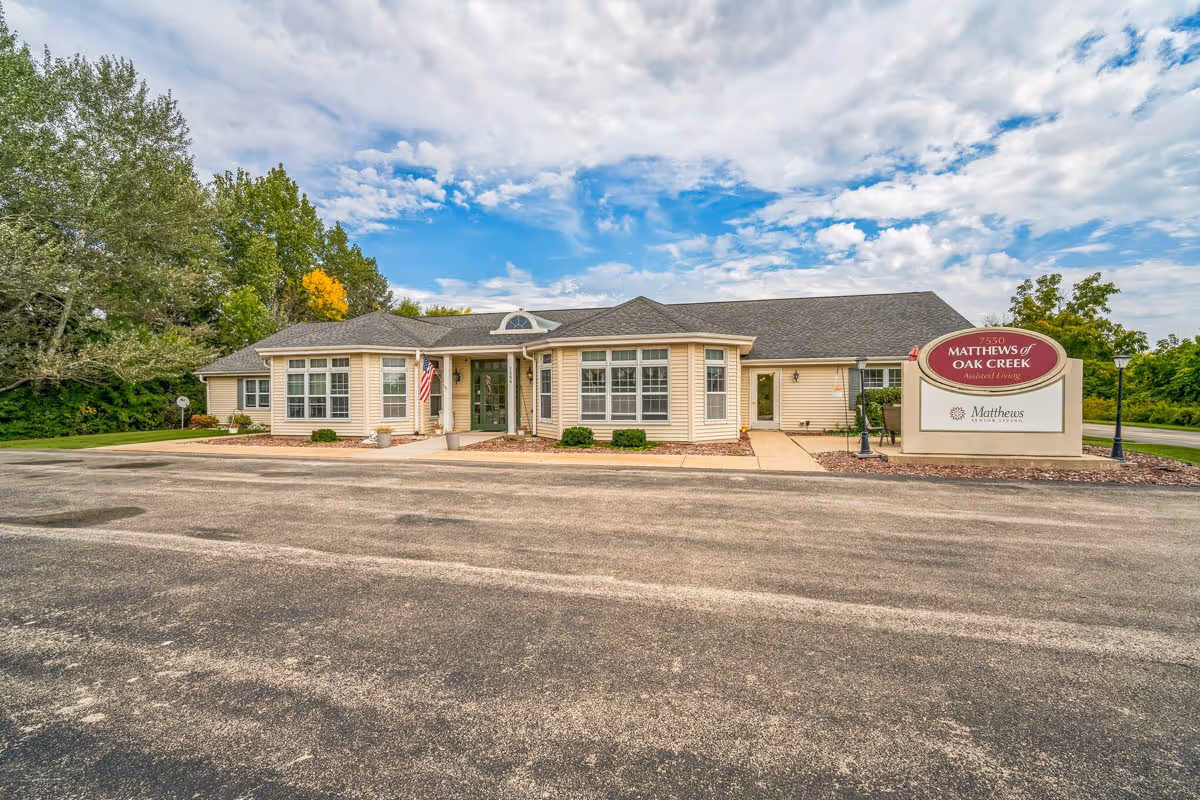 Exterior view of a single-story beige building with multiple windows and a dark shingled roof, surrounded by greenery and trees under a partly cloudy sky. There is a paved parking area in front and a sign on the right side that reads 'Matthews of Oak Creek Assisted Living.'