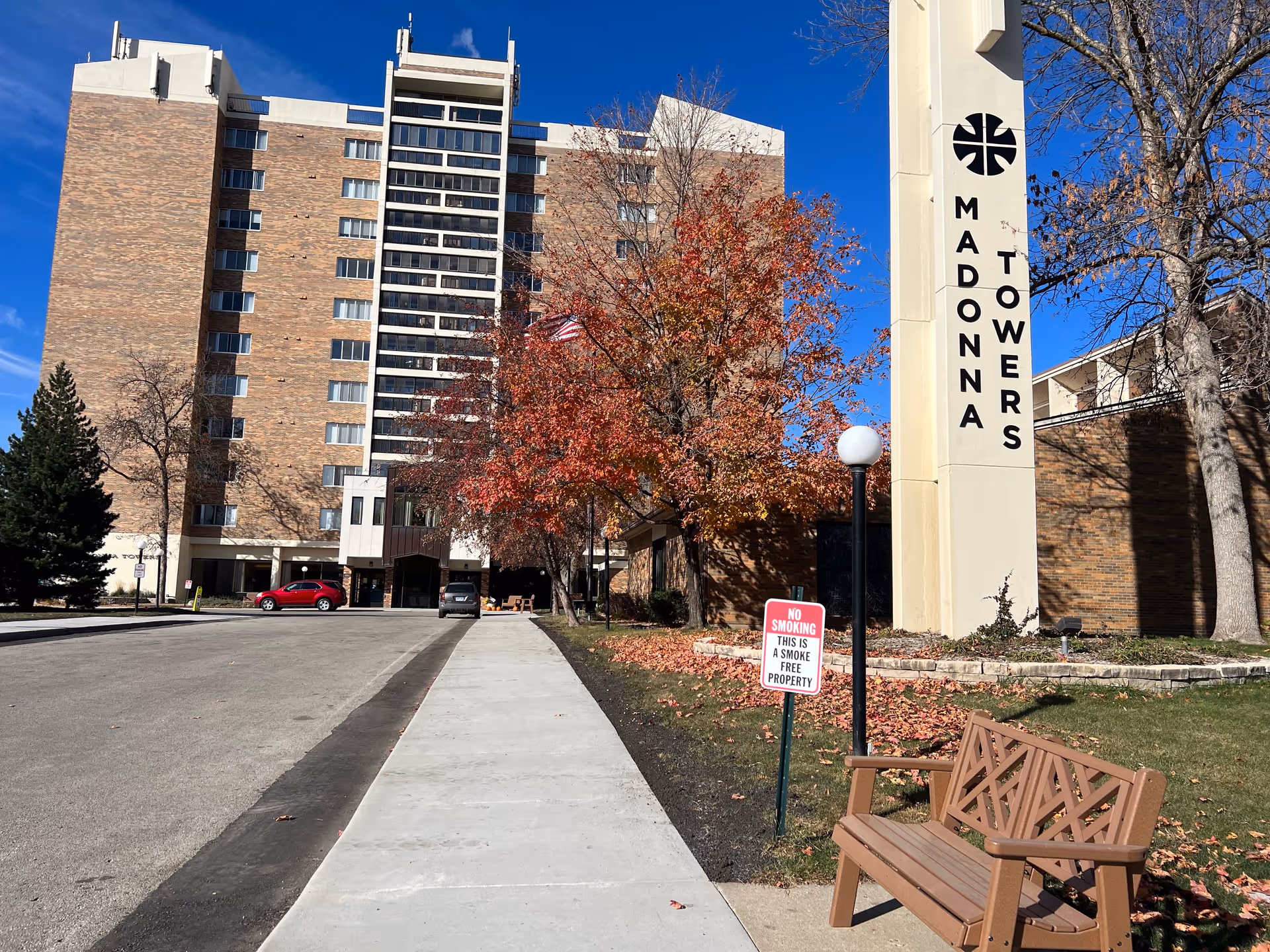 Tall brick Madonna Towers building with vertical sign, sidewalk, and a wooden bench.