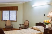 A cozy bedroom with a bed, nightstand, lamp, flowers, and two chairs by a window.