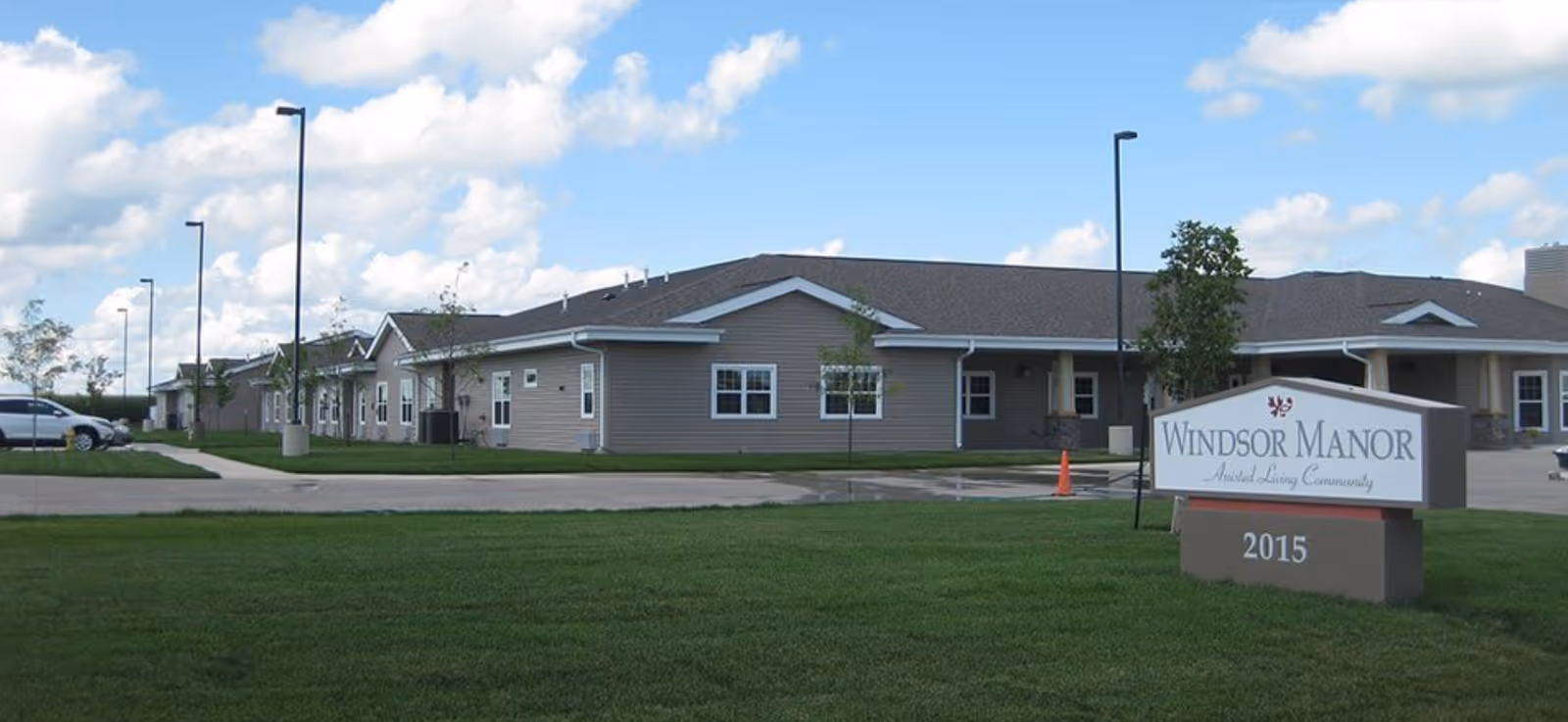 Exterior view of a single-story assisted living community building with beige siding and a dark roof under a partly cloudy blue sky. There is a sign in front that reads 'Windsor Manor Assisted Living Community 2015'. The area has green grass, a paved driveway, and several street lamps.