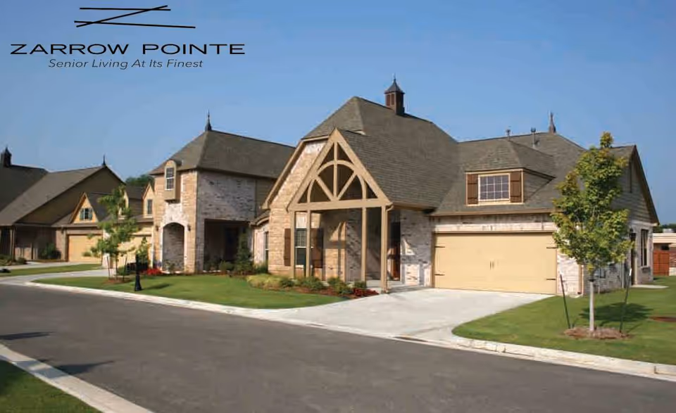 Exterior view of a senior living facility named Zarrow Pointe, showing multiple single-story houses with brick and wood accents, garages, manicured lawns, and a clear blue sky.