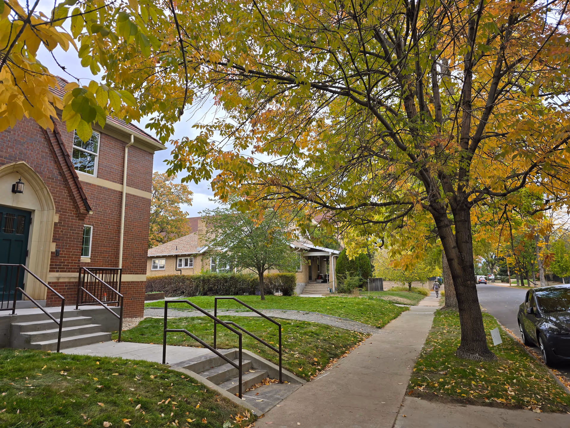 Sidewalk and street lined with autumn trees beside the front of a brick building and nearby houses.