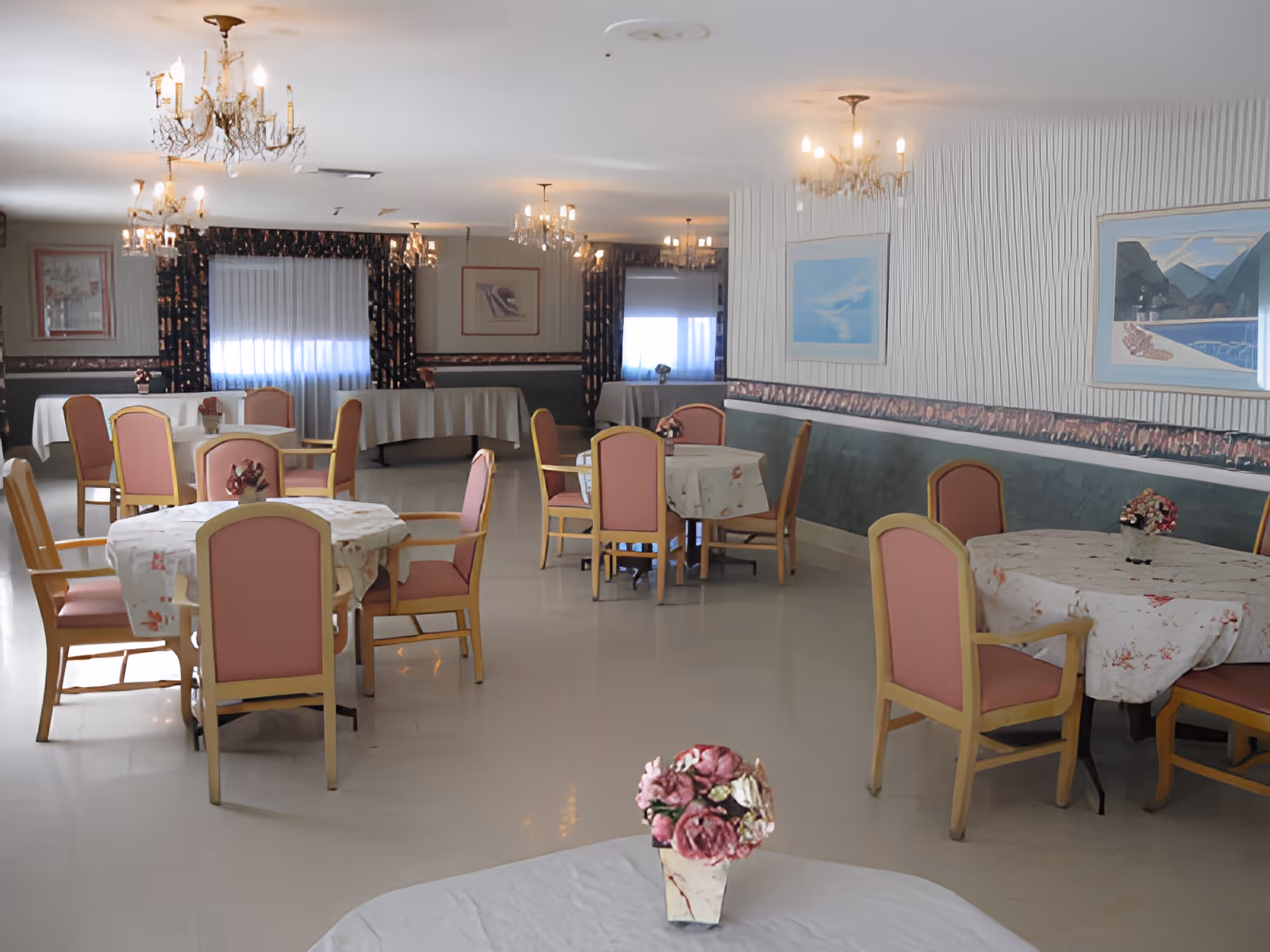 A dining room with several round tables covered with white tablecloths and pink cushioned chairs. Each table has a small floral centerpiece. The room is decorated with chandeliers, framed artwork on the walls, and windows with curtains.