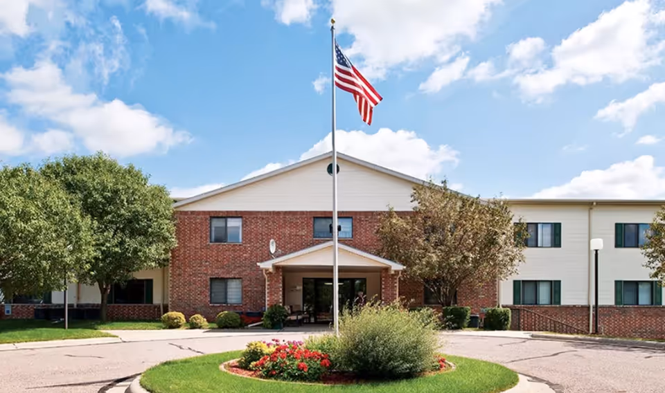 Front exterior view of The Heritage at Northern Hills building with a circular driveway, a flagpole with an American flag, green trees, and a flower bed under a partly cloudy sky.