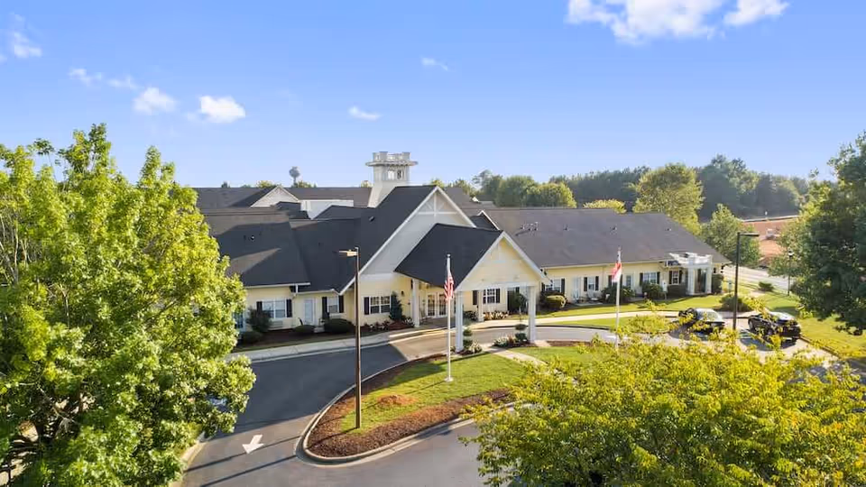 Front exterior of a single-story senior living facility with a covered porte-cochere, circular driveway, flagpoles, and surrounding trees.