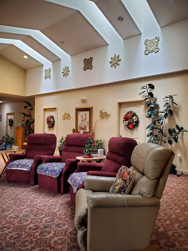A cozy living room area in a senior living facility with four recliner chairs, three in burgundy with floral covers and one beige with a decorative pillow. The room has a patterned carpet, potted plants, and doors decorated with holiday wreaths. The ceiling features skylights and decorative wall hangings.