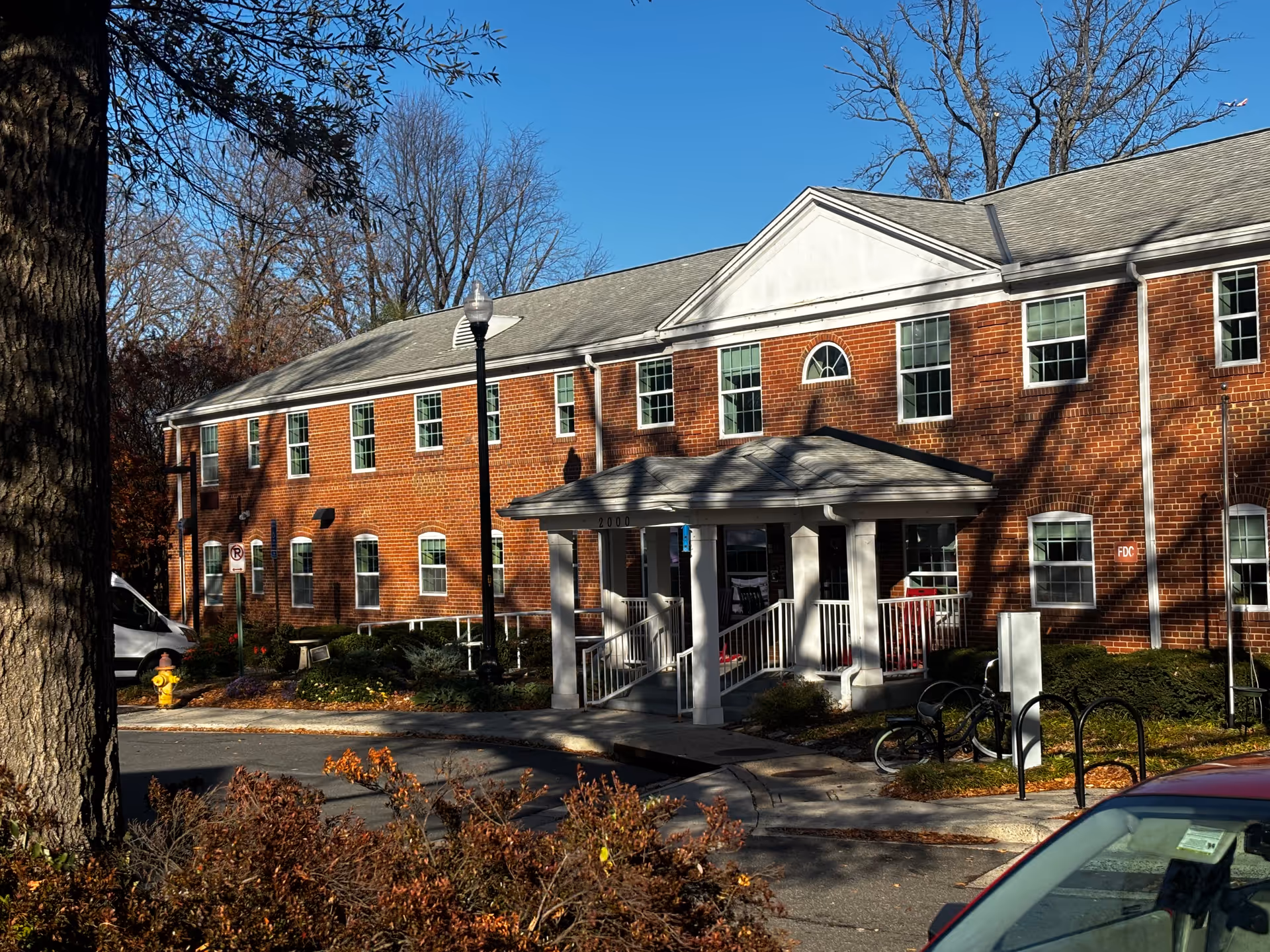 Two-story red brick building with a covered entrance, multiple windows, and trees in front under a clear blue sky.
