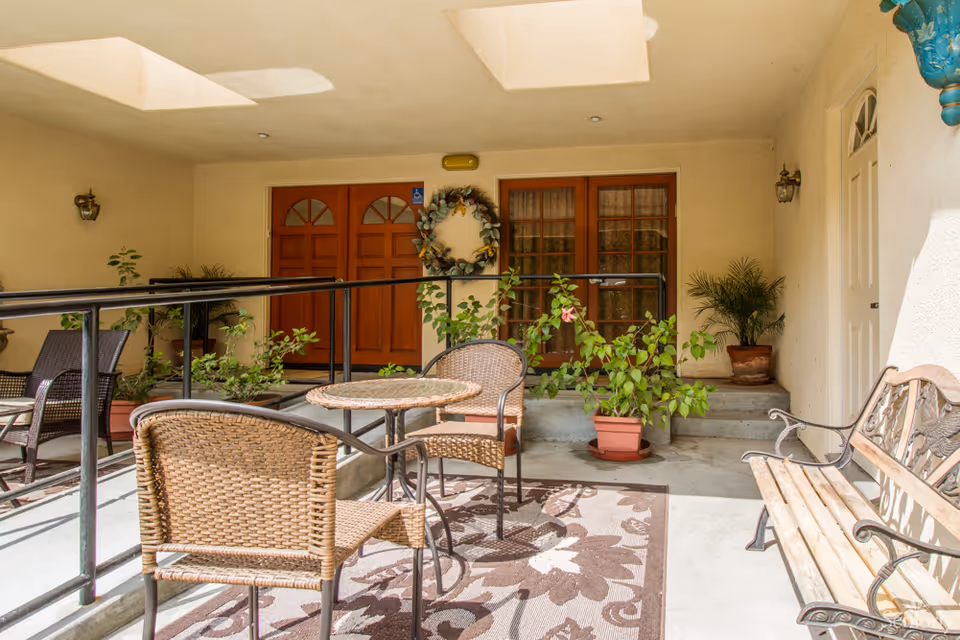 A covered outdoor patio area with a small round table and two wicker chairs on a floral patterned rug. There is a wooden bench with metal armrests on the right side. Several potted plants are placed around the space, including near two wooden doors and a white door. A decorative wreath hangs on the wall between the two wooden doors. The area has a beige wall and ceiling with recessed lighting and a skylight.