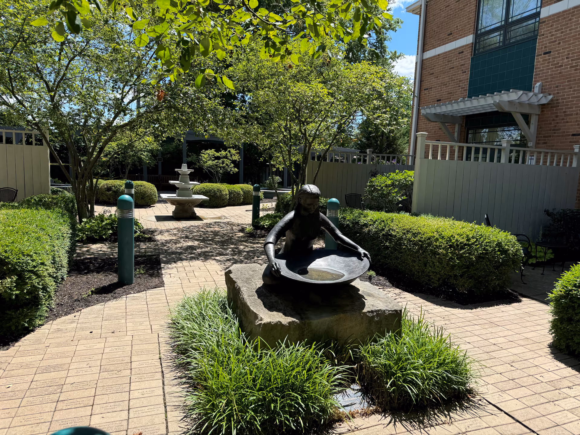 Sunlit outdoor courtyard with a fountain sculpture, greenery, and seating near a brick building.